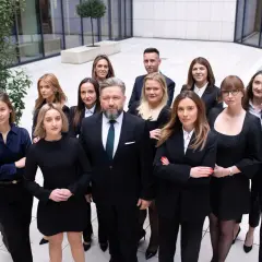 Grupa prawników w garniturach i sukienkach stoi przed nowoczesnym budynkiem.