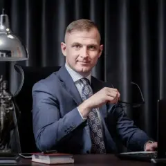 Mężczyzna w garniturze siedzi przy biurku, trzymając okulary. Obok statua Temidy.