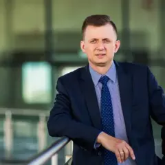 Mężczyzna w garniturze i krawacie opiera się o barierkę na tle nowoczesnego budynku.
