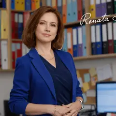 Kobieta w niebieskiej marynarce stoi przed regałami z kolorowymi segregatorami.