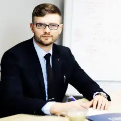 Mężczyzna w garniturze i okularach siedzi przy biurku, pisząc.