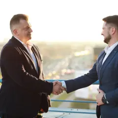 Dwóch mężczyzn w garniturach ściska sobie dłonie na tle miasta.