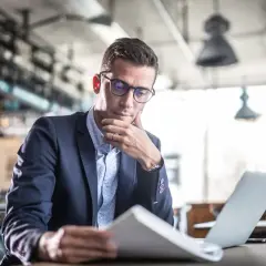 Mężczyzna w okularach i garniturze analizuje dokumenty przy laptopie w nowoczesnym wnętrzu.