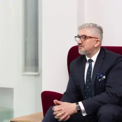 Mężczyzna w garniturze siedzi na czerwonym fotelu, patrząc w bok.