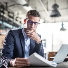 Mężczyzna w okularach i garniturze analizuje dokumenty przy laptopie w kawiarni.