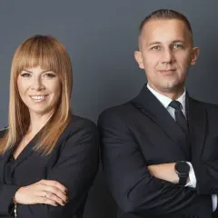 Kobieta i mężczyzna w czarnych garniturach, stojący obok siebie ze skrzyżowanymi ramionami.