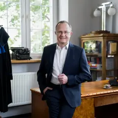 Mężczyzna w garniturze stoi w gabinecie obok biurka, na którym stoi komputer i filiżanka.