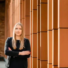 Kobieta w czarnym garniturze stoi ze skrzyżowanymi rękami na tle pomarańczowej fasady.