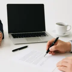 Podpisanie umowy z członkiem zarządu. Dwie osoby w garniturach przy biurku, jedna podpisuje dokument, obok laptop i kawa.