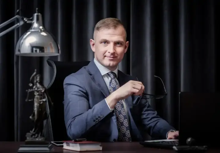 Mężczyzna w garniturze siedzi przy biurku, trzymając okulary. Obok statua Temidy.