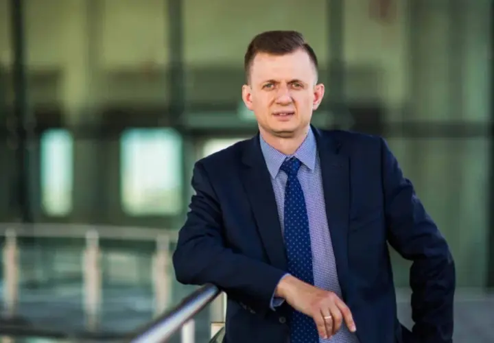 Mężczyzna w garniturze i krawacie opiera się o barierkę na tle nowoczesnego budynku.