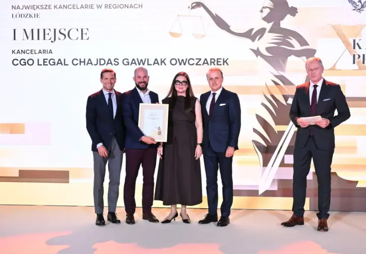 Pięć osób w garniturach i sukience stoi na scenie, odbierając nagrodę.