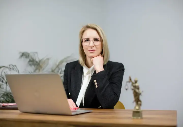 Kobieta w okularach i żakiecie siedzi przy biurku z laptopem i statuetką Temidy.