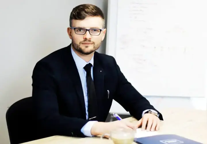 Mężczyzna w garniturze i okularach siedzi przy biurku, pisząc.
