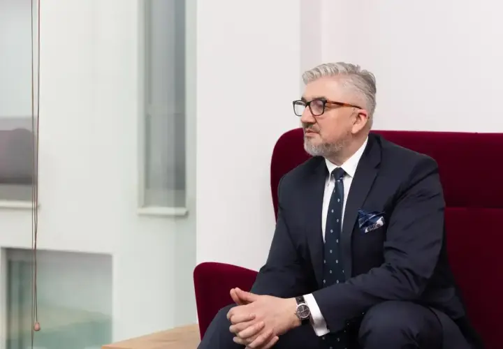 Mężczyzna w garniturze siedzi na czerwonym fotelu, patrząc w bok.