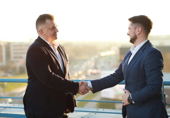 Dwóch mężczyzn w garniturach ściska sobie dłonie na tle miasta.