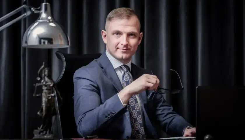 Mężczyzna w garniturze siedzi przy biurku, trzymając okulary. Obok statua Temidy.