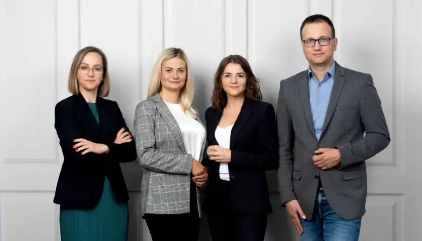 Cztery osoby w strojach biznesowych stoją przed jasną ścianą.