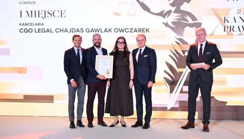Pięć osób w garniturach i sukience stoi na scenie, odbierając nagrodę.