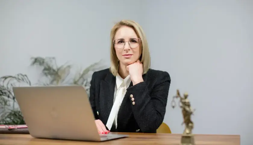 Kobieta w okularach i żakiecie siedzi przy biurku z laptopem i statuetką Temidy.