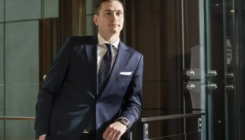 Mężczyzna w granatowym garniturze i krawacie stoi oparty o barierkę w nowoczesnym wnętrzu.