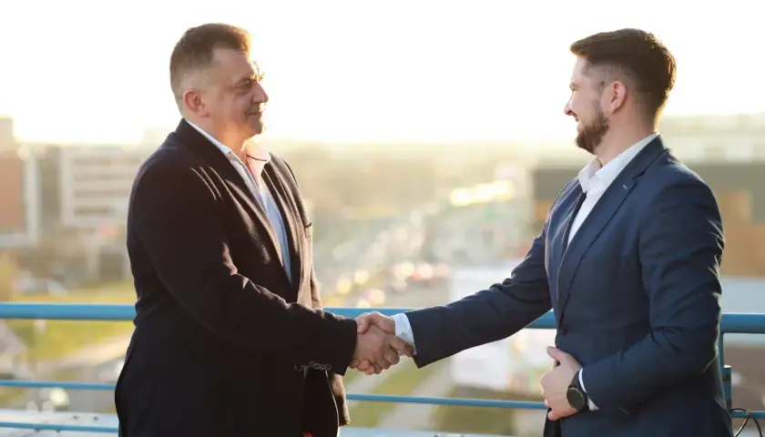 Dwóch mężczyzn w garniturach ściska sobie dłonie na tle miasta.