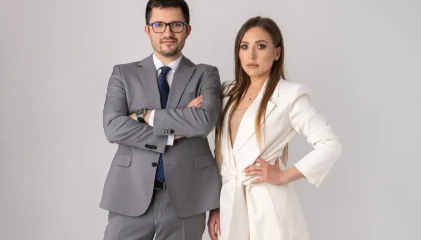 Para prawników w garniturach, mężczyzna w szarym, kobieta w białym, na jasnym tle.