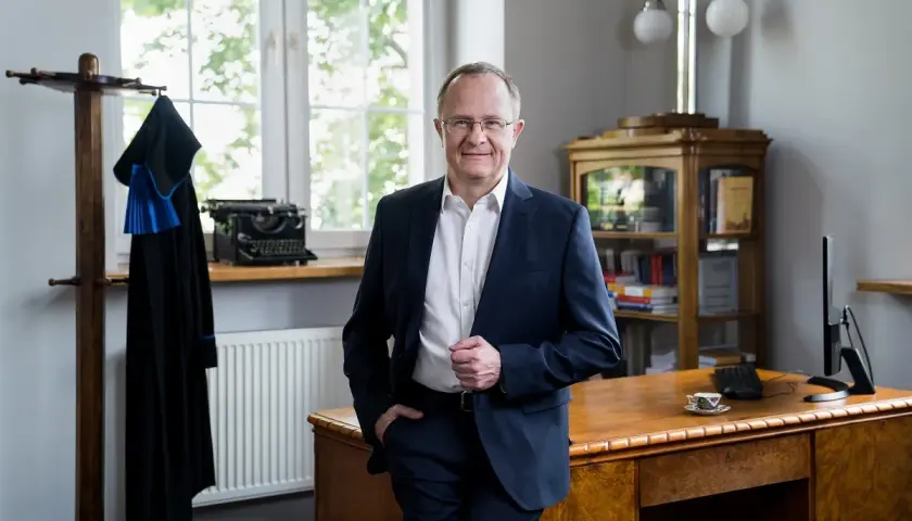 Mężczyzna w garniturze stoi w gabinecie obok biurka, na którym stoi komputer i filiżanka.