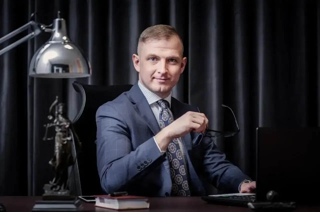 Mężczyzna w garniturze siedzi przy biurku, trzymając okulary. Obok statua Temidy.