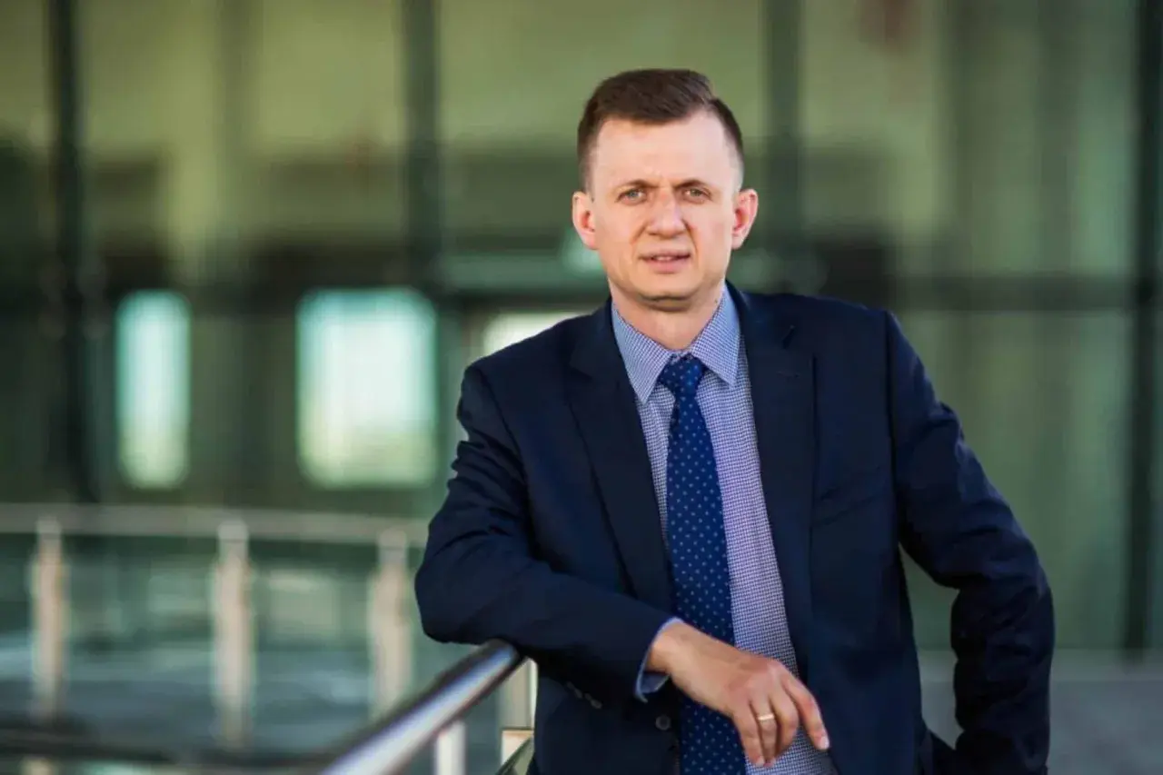Mężczyzna w garniturze i krawacie opiera się o barierkę na tle nowoczesnego budynku.