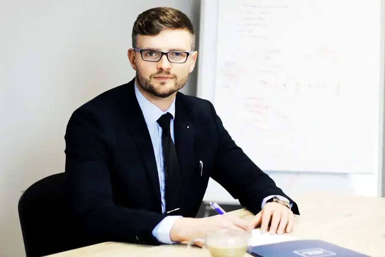 Mężczyzna w garniturze i okularach siedzi przy biurku, pisząc.
