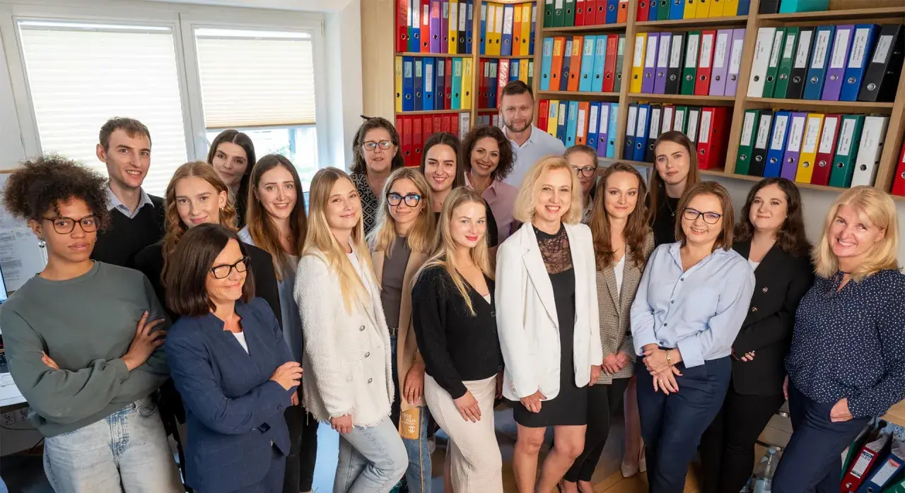Grupa uśmiechniętych doradców podatkowych w biurze, na półkach kolorowe segregatory.