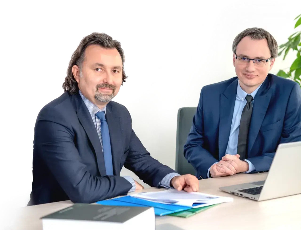 Dwóch mężczyzn w garniturach siedzi przy biurku z dokumentami i laptopem.