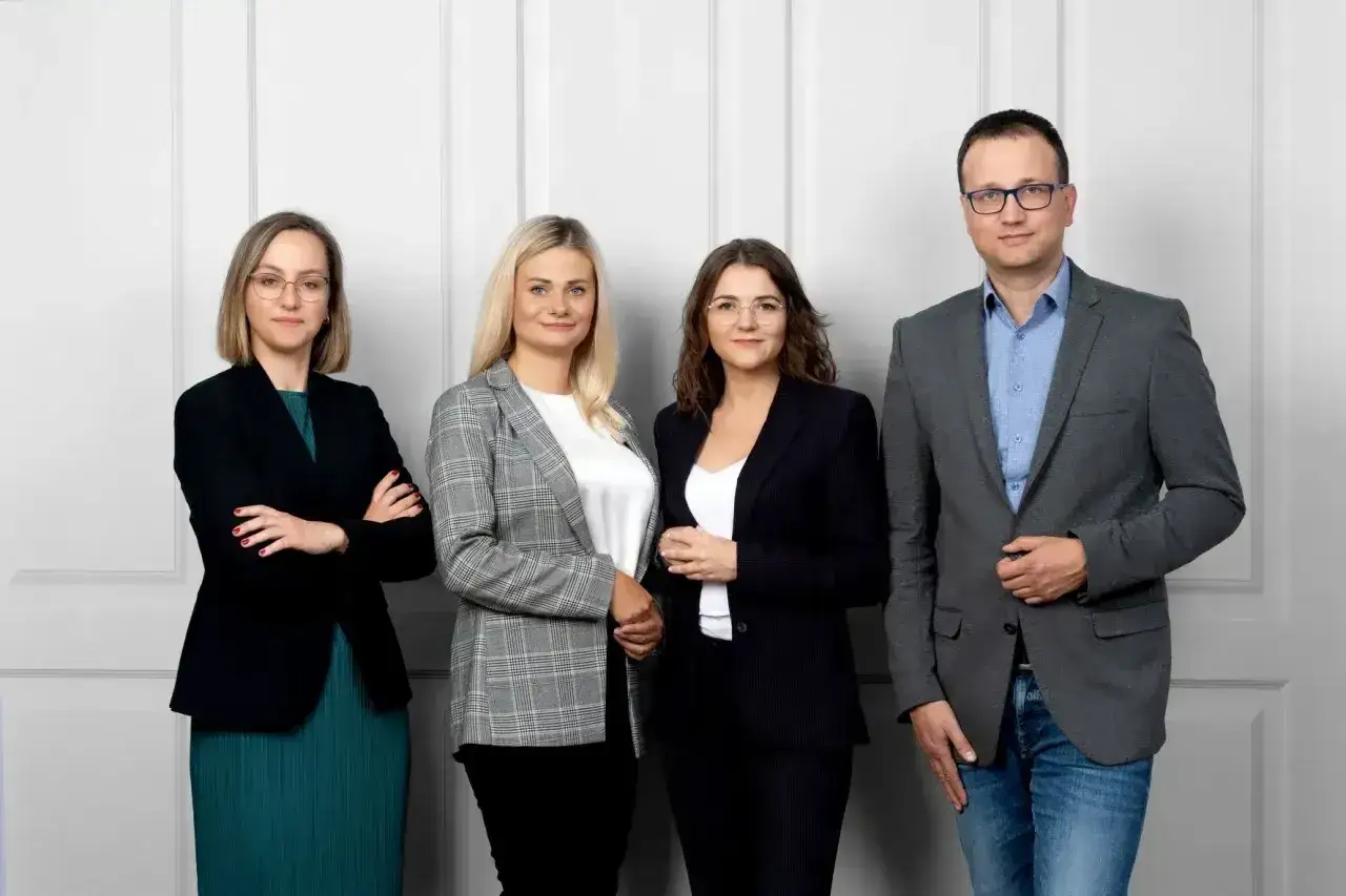 Cztery osoby w strojach biznesowych stoją przed jasną ścianą.