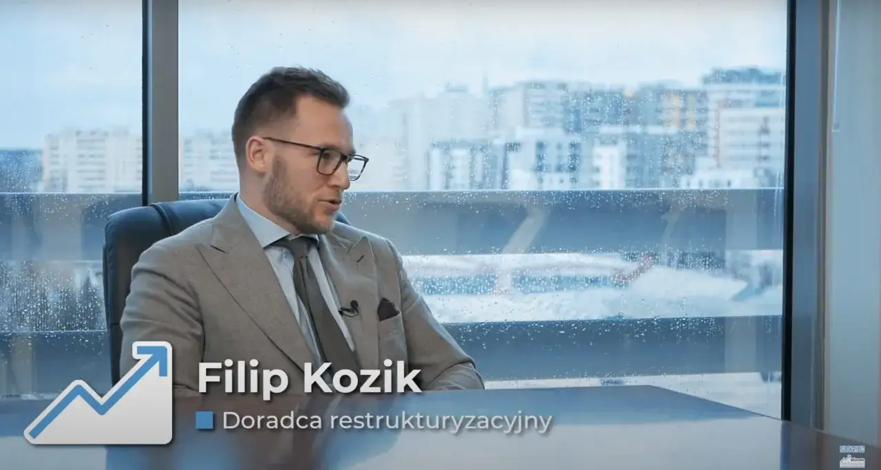Filip Kozik, doradca restrukturyzacyjny, w garniturze siedzi przy biurku. W tle okno z kroplami deszczu i widokiem na miasto.