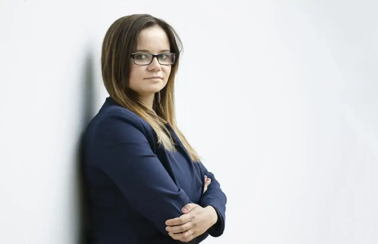 Kobieta w okularach i granatowej marynarce, oparta o ścianę, ze skrzyżowanymi rękami.