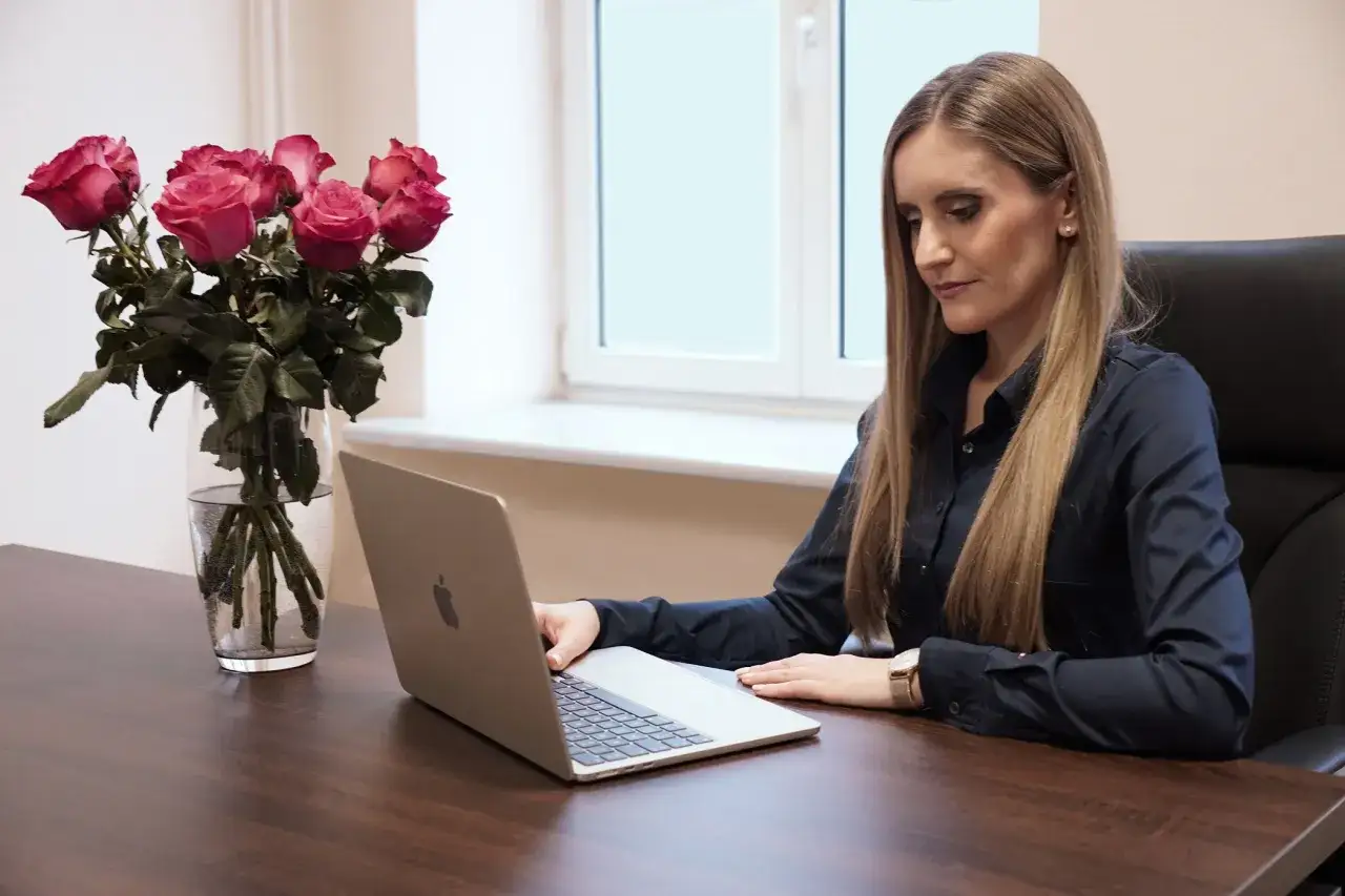 Kobieta w czarnej koszuli pracuje przy laptopie, obok bukiet róż w wazonie.