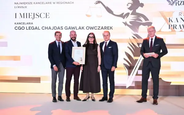 Pięć osób w garniturach i sukience stoi na scenie, odbierając nagrodę.
